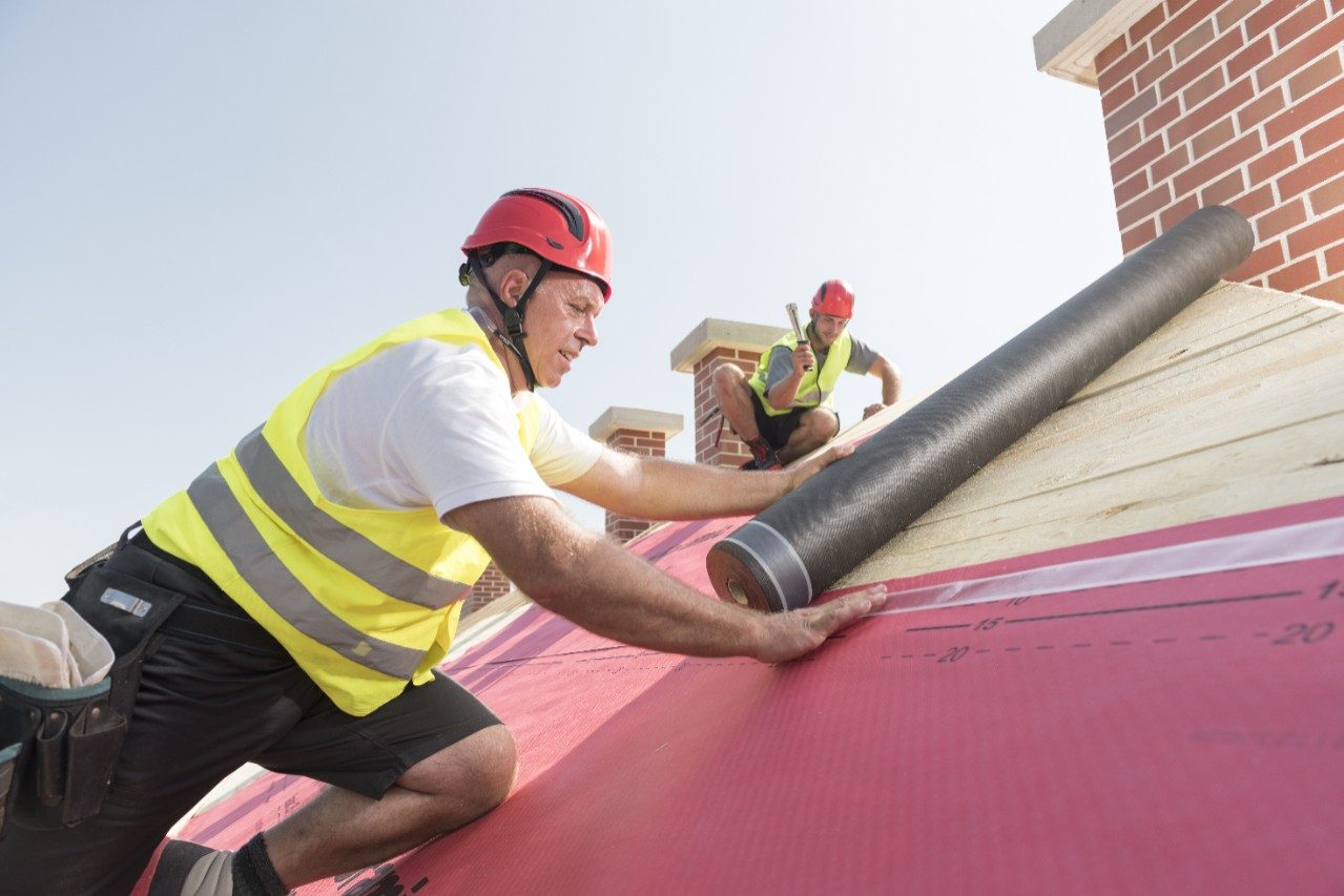 Urban roofers applying roof underlay sheet wearing hard hat and safety jacket brick chimney