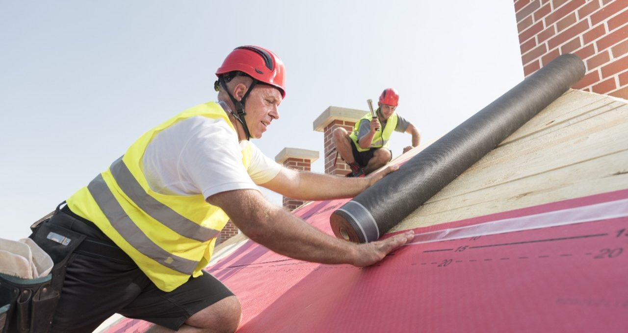 Urban roofers applying roof underlay sheet wearing hard hat and safety jacket brick chimney