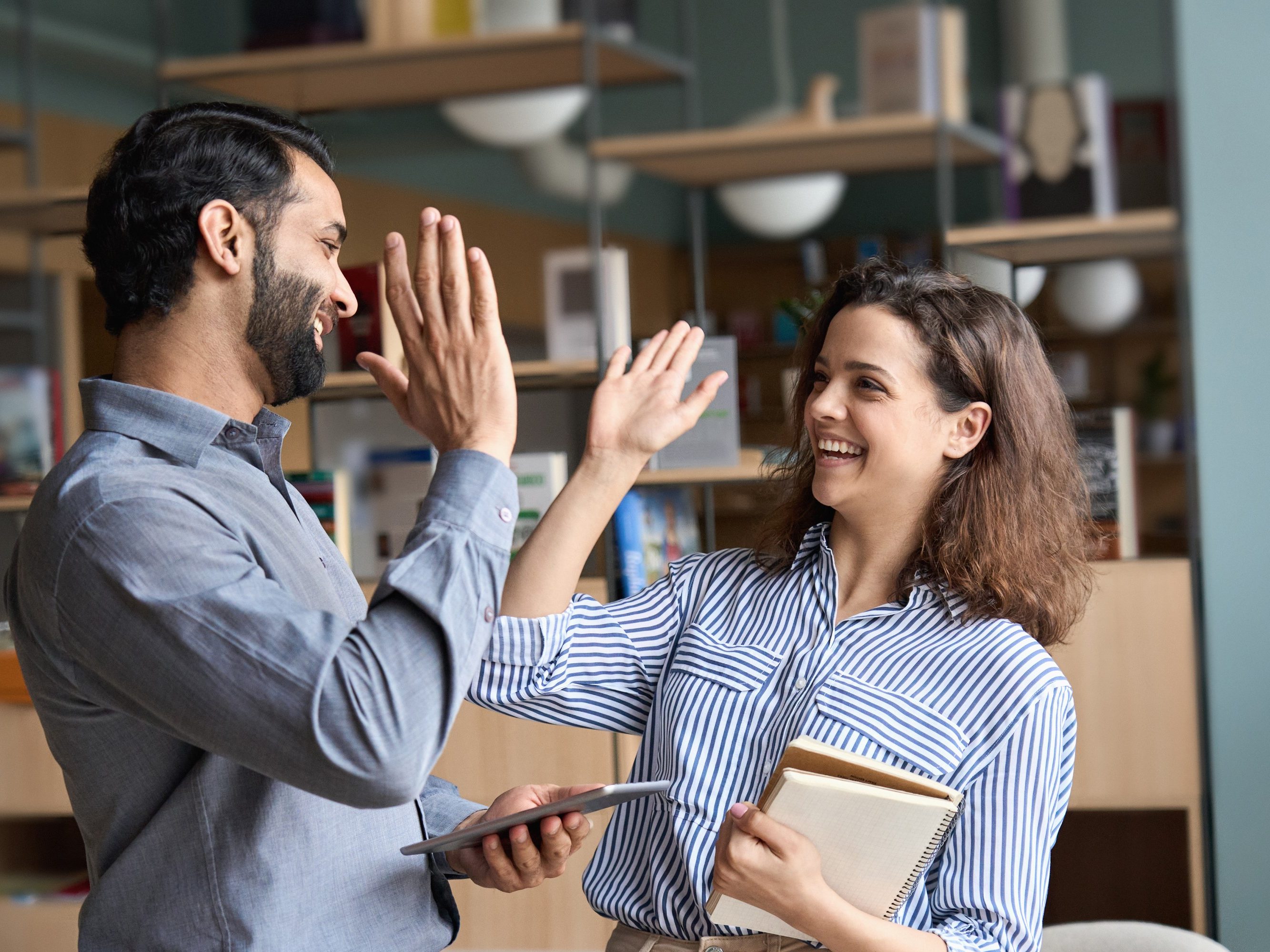 Two happy friendly diverse professionals, teacher and student giving high five standing in office celebrating success, good cooperation result, partnership teamwork and team motivation in office work.