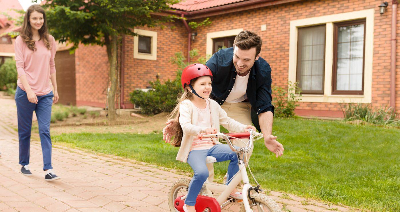 Father protecting little girl while learning to ride the bike while mother watches them