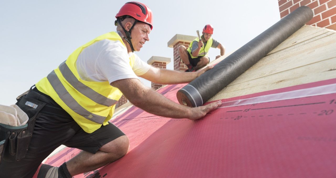 Urban roofers applying roof underlay sheet wearing hard hat and safety jacket brick chimney